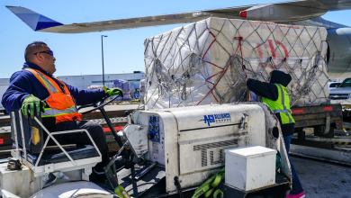 A man with an orange vest drives a cargo tug up to an aircraft to load a pallet.