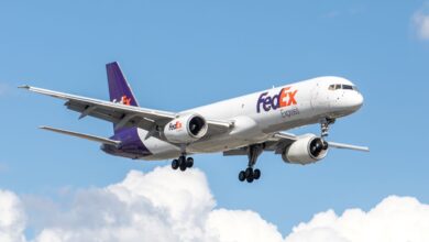 Purple-tail FedEx jet with wheels down approaches airport on a bright sunny day with light clouds in the background.