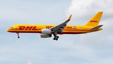 A mustard-yellow plane with red DHL lettering flies with wheels down.