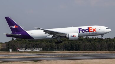 A FedEx plane with purple tail lands on runway with trees in background.