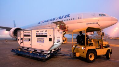 Ground view of air cargo containers pulled by an airport tractor to the side of a light-blue Korean Air jet.