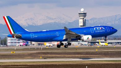 A bright blue ITA Airways plane takes off with mountains in the background.