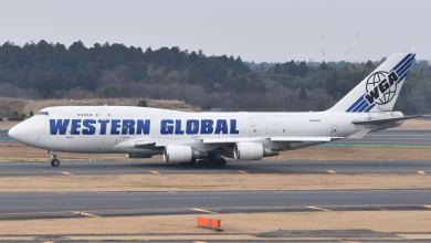 A white 747 jumbo jet with blue lettering: Western Global rolls down taxiway on a hazy day.