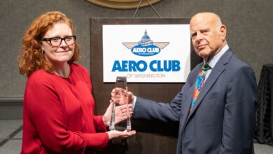 A white woman hands an award to an older white man in front of an Aero Club sign.