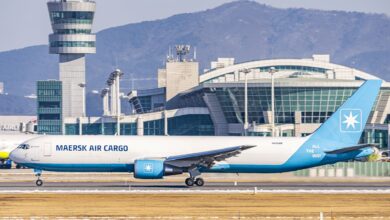A light-blue tailed Maersk Air Cargo jet on the taxiway with an airport control tower and passenger terminal in the background.