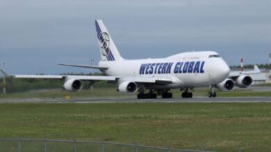 A white jumbo jet with dark blue lettering "Western Global" rolls on taxiway with grass on each side.