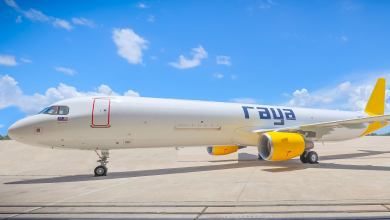 A yellow-tailed jet with yellow engines and "Raya" lettering sits on the tarmac under a bright blue sky in a close up view from the side.