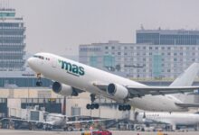 A white "mas" cargo jet takes off from Los Angeles airport with buildings in the background on a gray day.