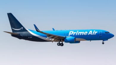 An all-blue Prime Air freighter with wheels down approaches an airport against a blue sky.