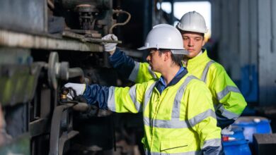 Two people wearing hard hats and protective jackets look at a train wheel.