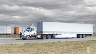 A white tractor-trailer on the highway near a train hauling cars