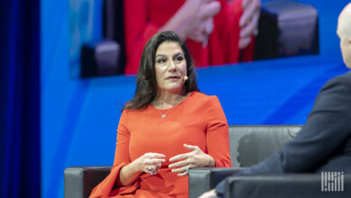 Woman with brown hair and red dress in a chair discussing issues on stage against a blue background.