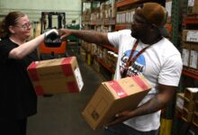 A white woman and a black man bump fists holding a package in in a storage room of a factory.