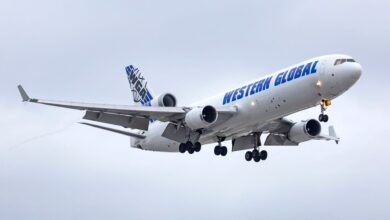 A large tri-engine Western Global jet with blue lettering with wheels down on approach to an airport.