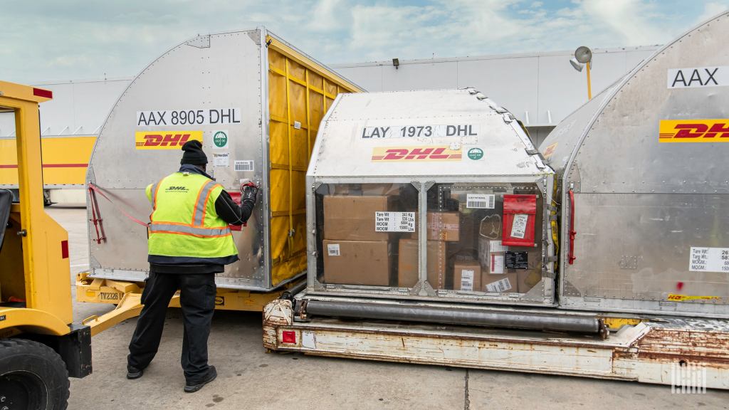 A DHL worker with a yellow vest arranges air containers on the tarmac.