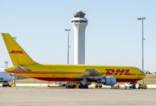 A large yellow DHL jet sits on the tarmac with an airport control tower in the background.