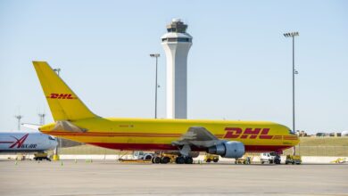 A large yellow DHL jet sits on the tarmac with an airport control tower in the background.