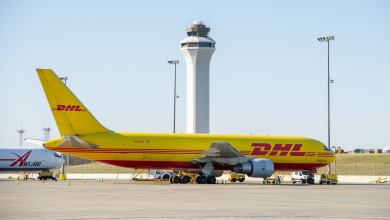 A large yellow DHL jet sits on the tarmac with an airport control tower in the background.