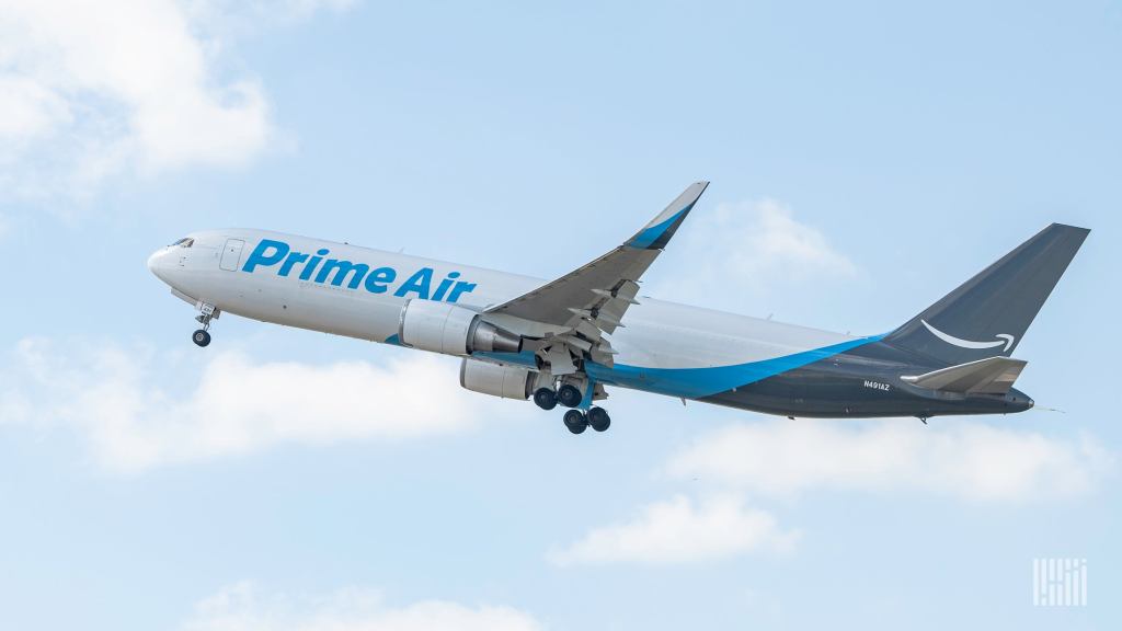 A Prime Air cargo jet with light blue lettering and dark blue tail takes off into blue sky with white clouds.