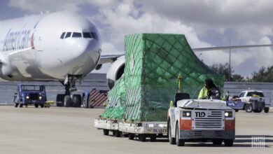 An airport tug pulls a flat trailer with a container draped in green plastic.