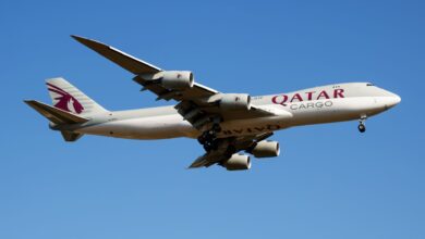 A gray Qatar Airways jumbo cargo jet seen from below as it flies low against a blue sky.