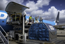 A light blue Amazon Prime plane on the ground with its side cago door open and a container ready to be loaded from the ground.