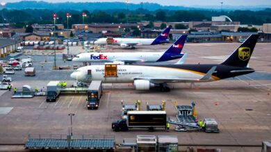 Purple-tail FedEx jets and a brown-tail UPS jet side by side with cargo containers and tractors at an airport.