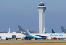 Amazon Prime Air freighters with blue accents on the tarmac underneath tall air traffic control tower.