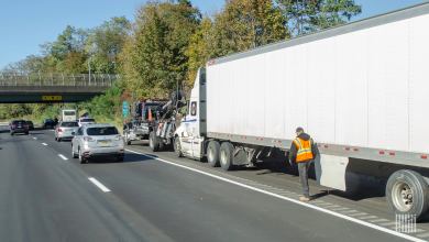 Semi-truck on the side of the highway with a driver in an orange vest walking next to it.