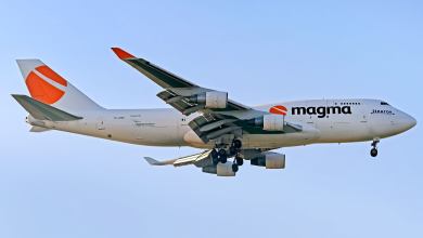 A Magma Aviation jumbo jet freighter with orange lettering seen from below as it comes in for landing with wheels down.