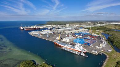 Image shows seaport berth, cargo vessel, sky, containers and shoreside structures.
