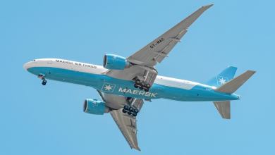 A Maersk Air Cargo jet with light-blue accents flies overhead with wheels down. View of belly.