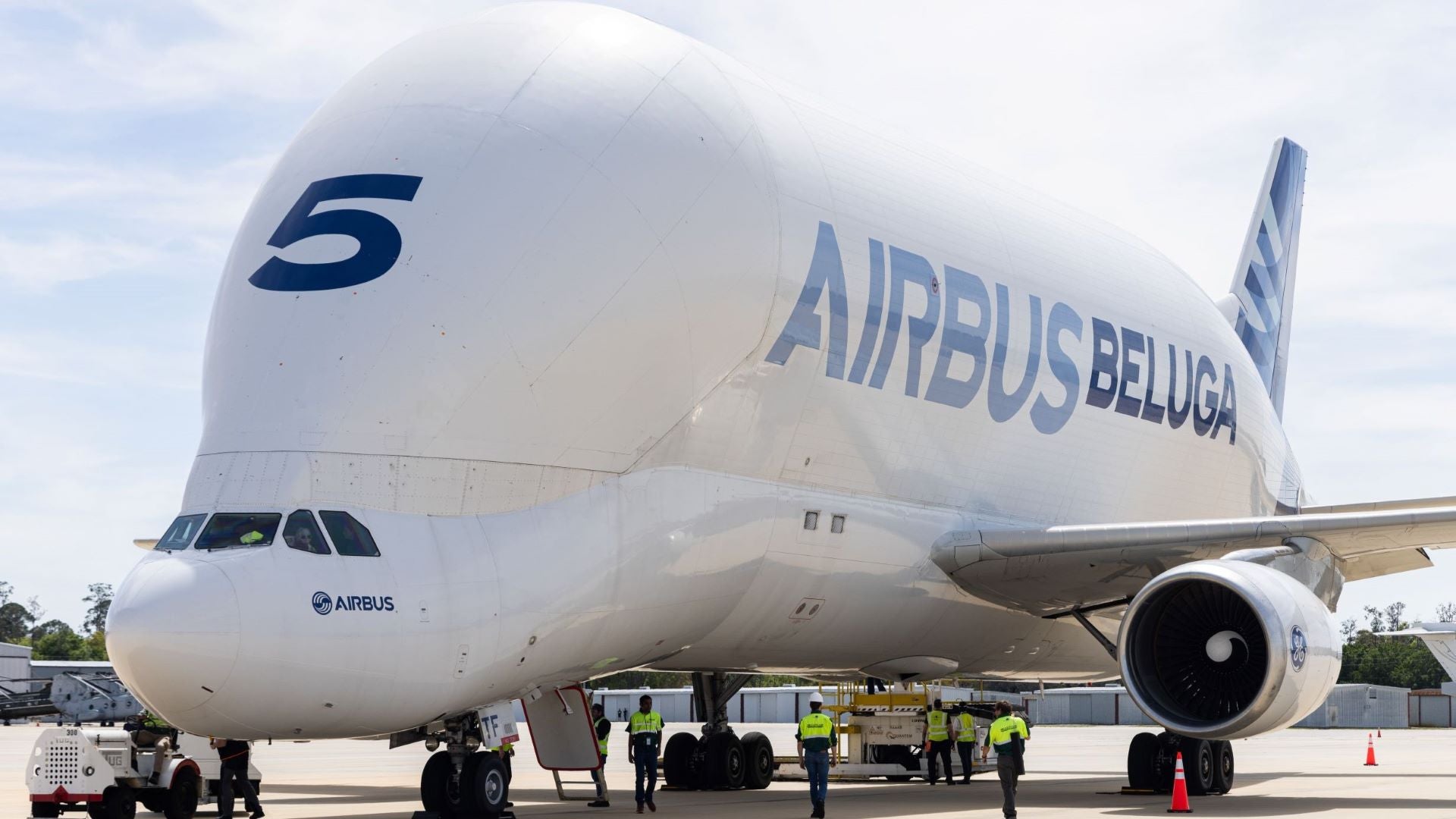 An Airbus Beluga freighter jet, with a bulbous fuselage, sits on the tarmac with workers milling about.