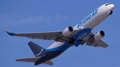 Large cargo jet with light blue markings and Amazon written on the bottom takes off in a blue sky, a seen from below.