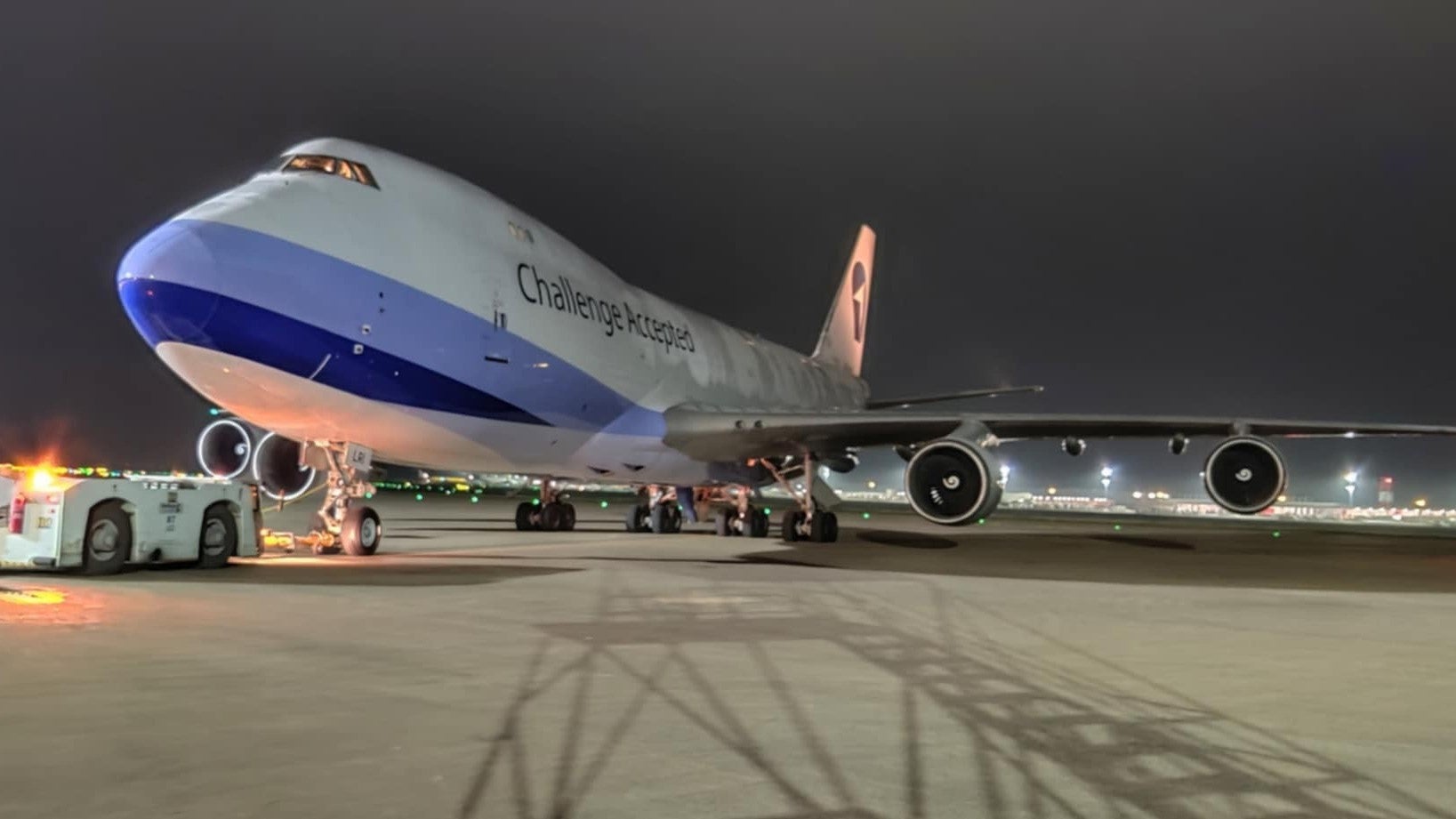 A jumbo cargo jet with blue accent paint sits on the tarmac at night, viewed from the left-front side.