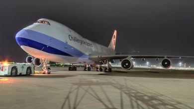 A jumbo cargo jet with blue accent paint sits on the tarmac at night, viewed from the left-front side.