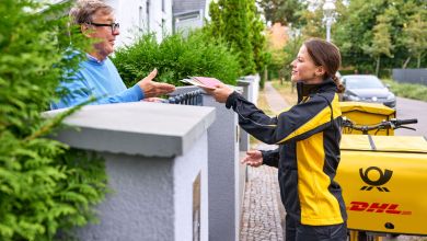 A female postal carrier with black hair wearing yellow Deutsche Post uniform hands mail to a home owner in Germany.