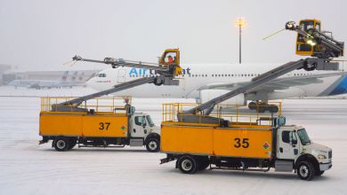 Orange deicing trucks drive on a snowy airfield with an airplane in the background.