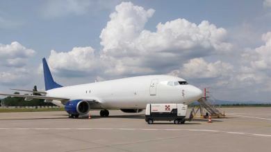 A blue-tailed cargo plane sits at an airport on a sunny day with white clouds in background.