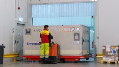 A man in a yellow-red uniform pushes a large container through a warehouse with the help of machinery.