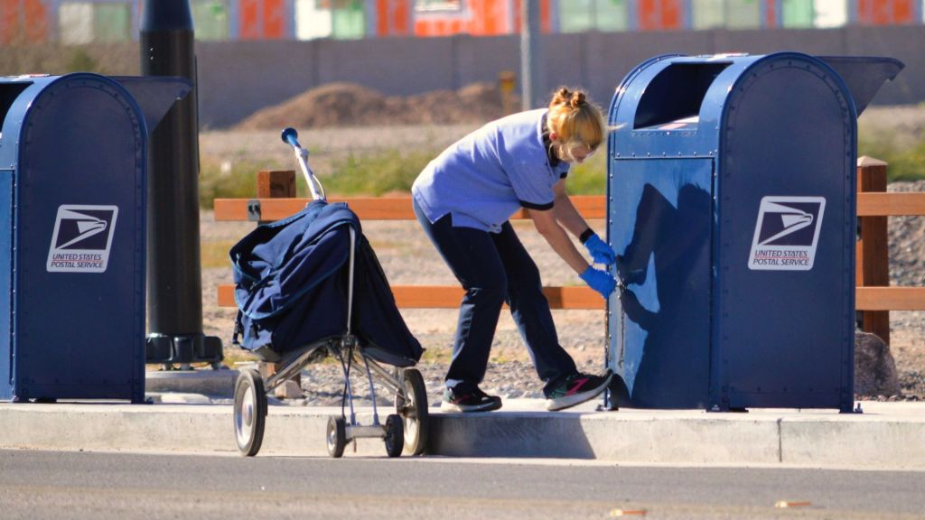 A female USPS worker bends to open a letter drop box and take the mail to the processing center.