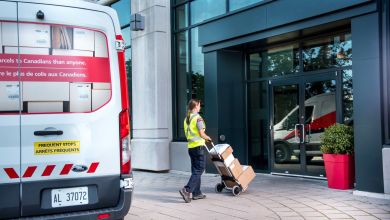 A mail carrier delivers packages from the van to an office building using a hand cart.