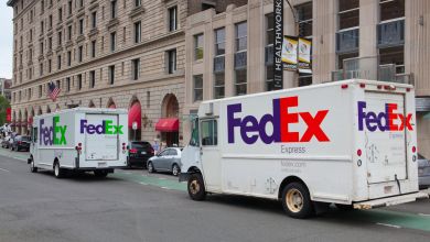 A FedEx Express truck with orange lettering and a FedEx Ground truck with green lettering parked together on a city street.