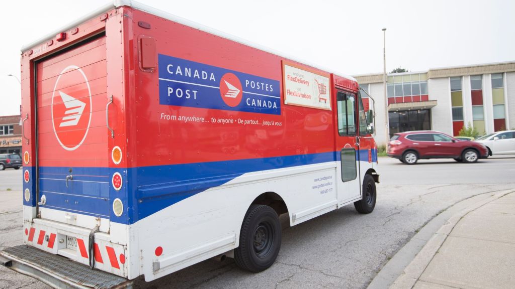 A red-blue-and-white Canada Post delivery vehicle, seen close up from the side rear on a roadway.