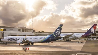 Alaska Airlines plane with a blue tail and Eskimo face painted sits beside a Hawaiian Airlines jet at an airport with a mix of sun and clouds.