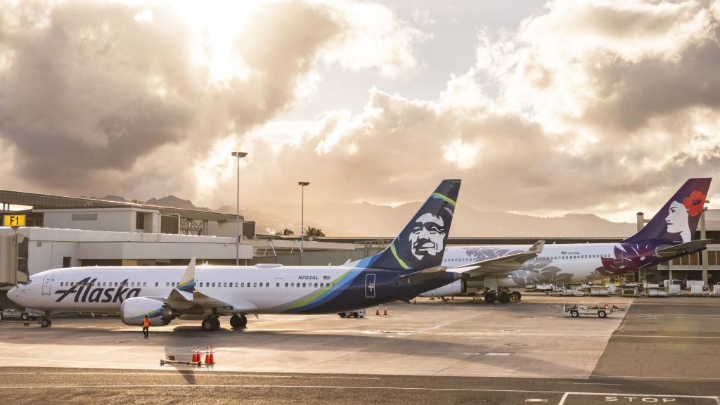 Alaska Airlines plane with a blue tail and Eskimo face painted sits beside a Hawaiian Airlines jet at an airport with a mix of sun and clouds.