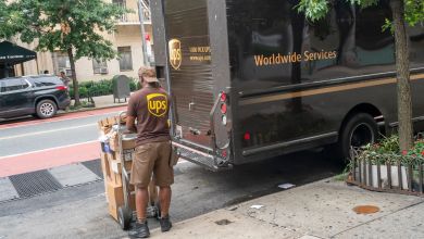 A UPS driver in a brown uniform unloads a brown UPS truck and puts packages on a hand cart in the city.