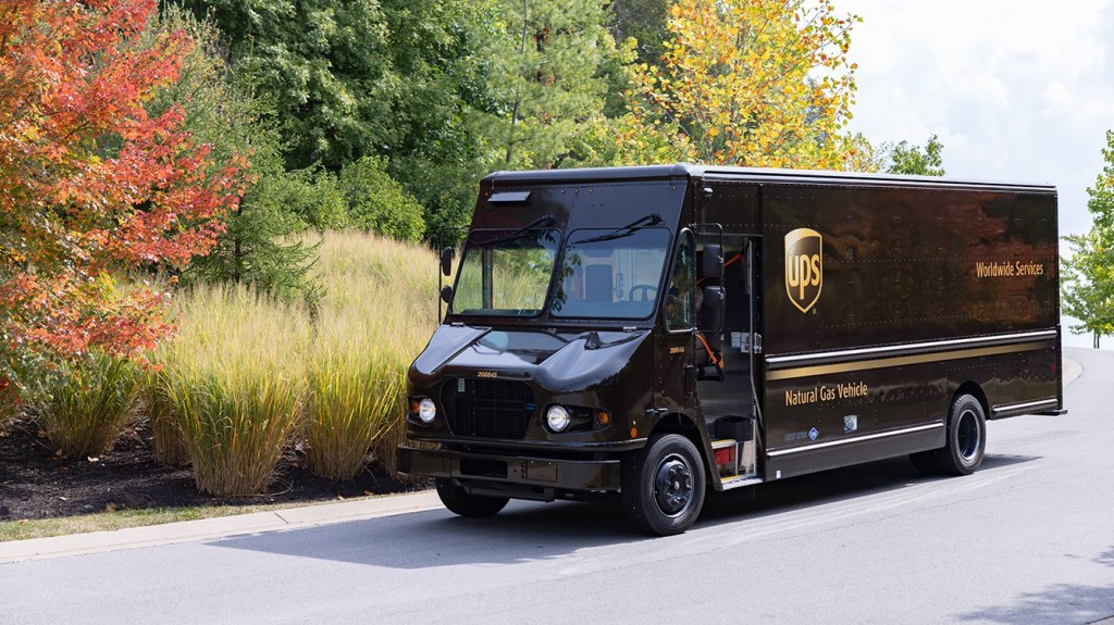 A brown UPS package car moves down a country road with tall grass and trees in the background.