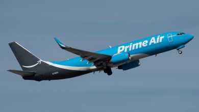 A two-toned blue Amazon Prime Air cargo jets takes off into a blue sky as seen from below.