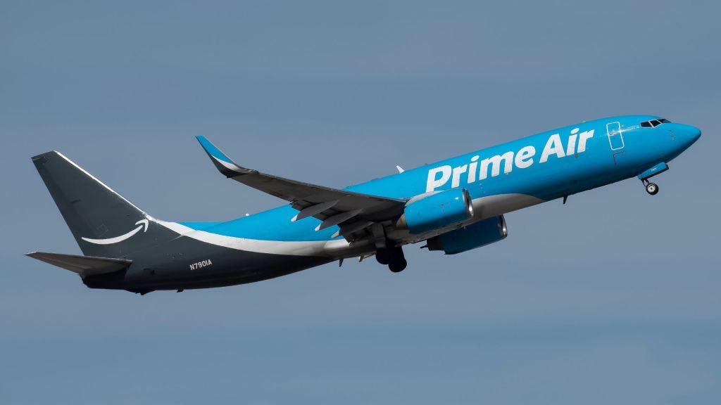 A two-toned blue Amazon Prime Air cargo jets takes off into a blue sky as seen from below.